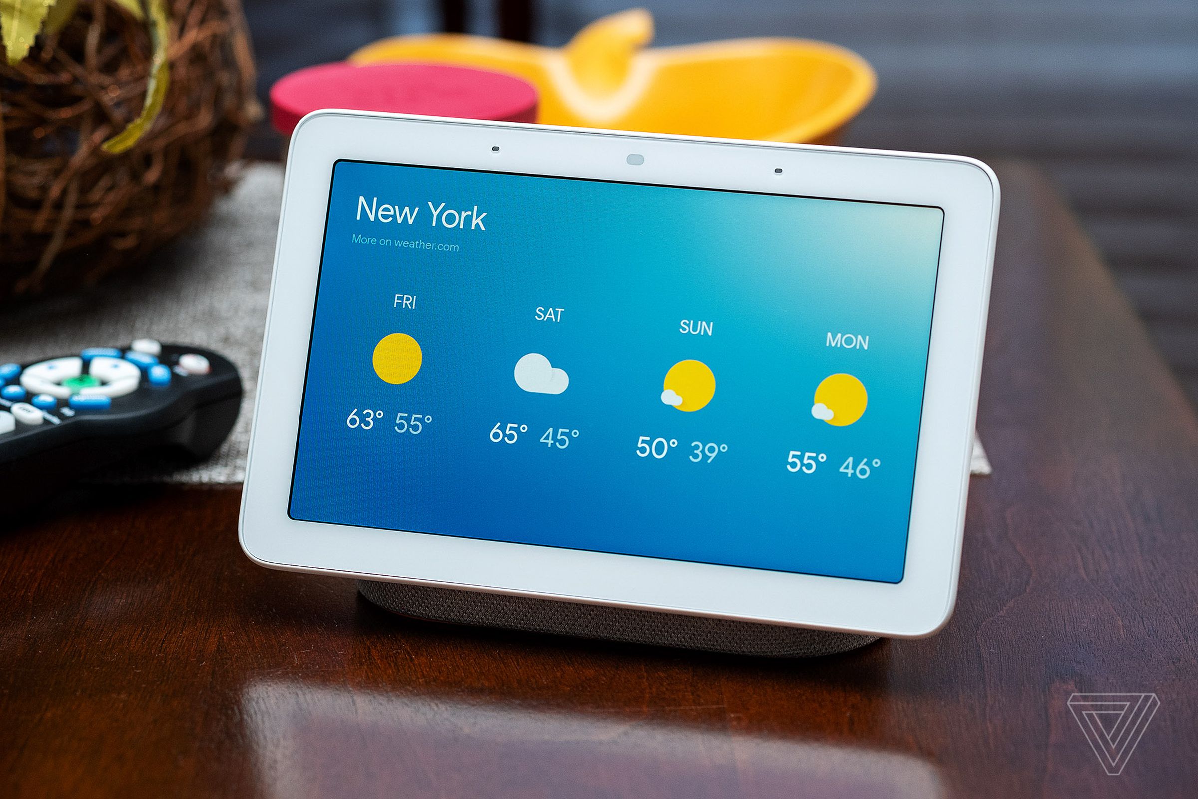 A first generation Google Nest Hub showing the weather, sitting on a desk.