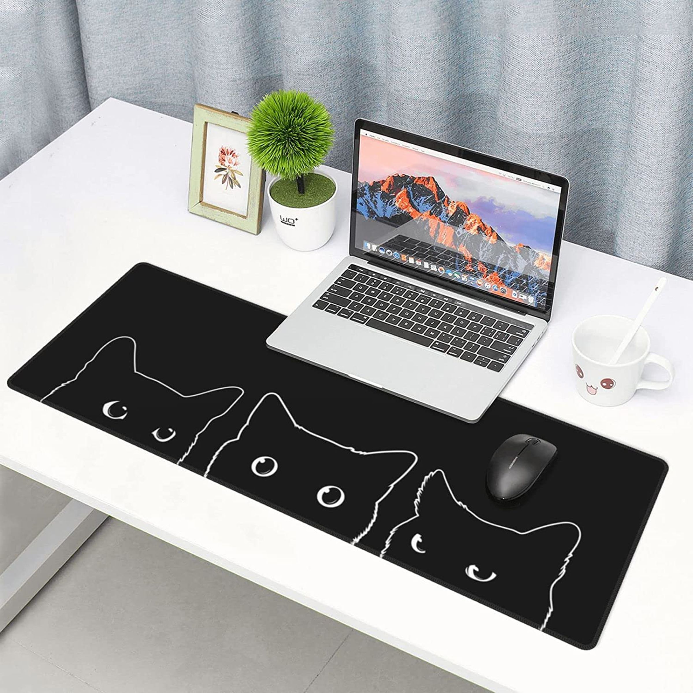 A long black mouse pad depicting the silhouette of three cats on a white desk with a laptop, a plant and a small framed photo.