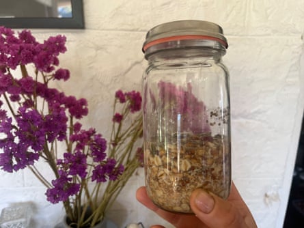 A hand holding a glass jar containing rolled oats and sunflower seeds against a white wall.  A jar of tall purple flowers is in the background.