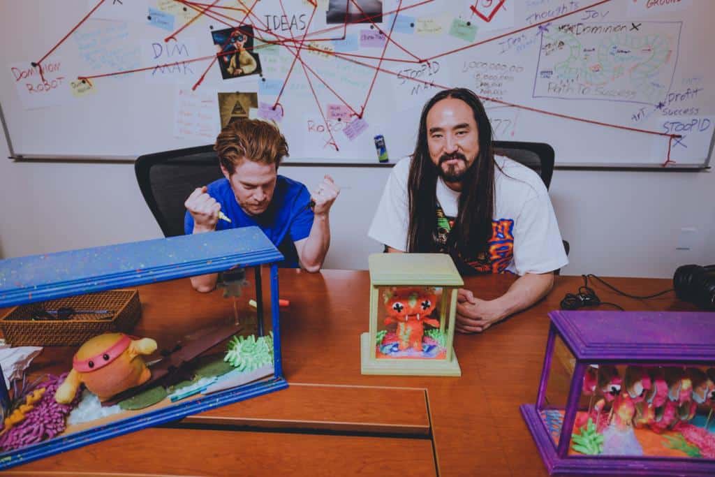 A photo of Steve Aoki and Seth Green, sitting at a desk with a physical copy of Aoki's Replicant X NFT project avatar.