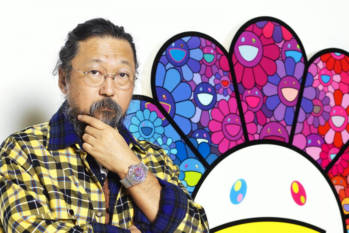 Takashi Murakami stands in front of a multicolored smiley face wearing a Hublot watch in support of the launch of Murakami X Hublot.