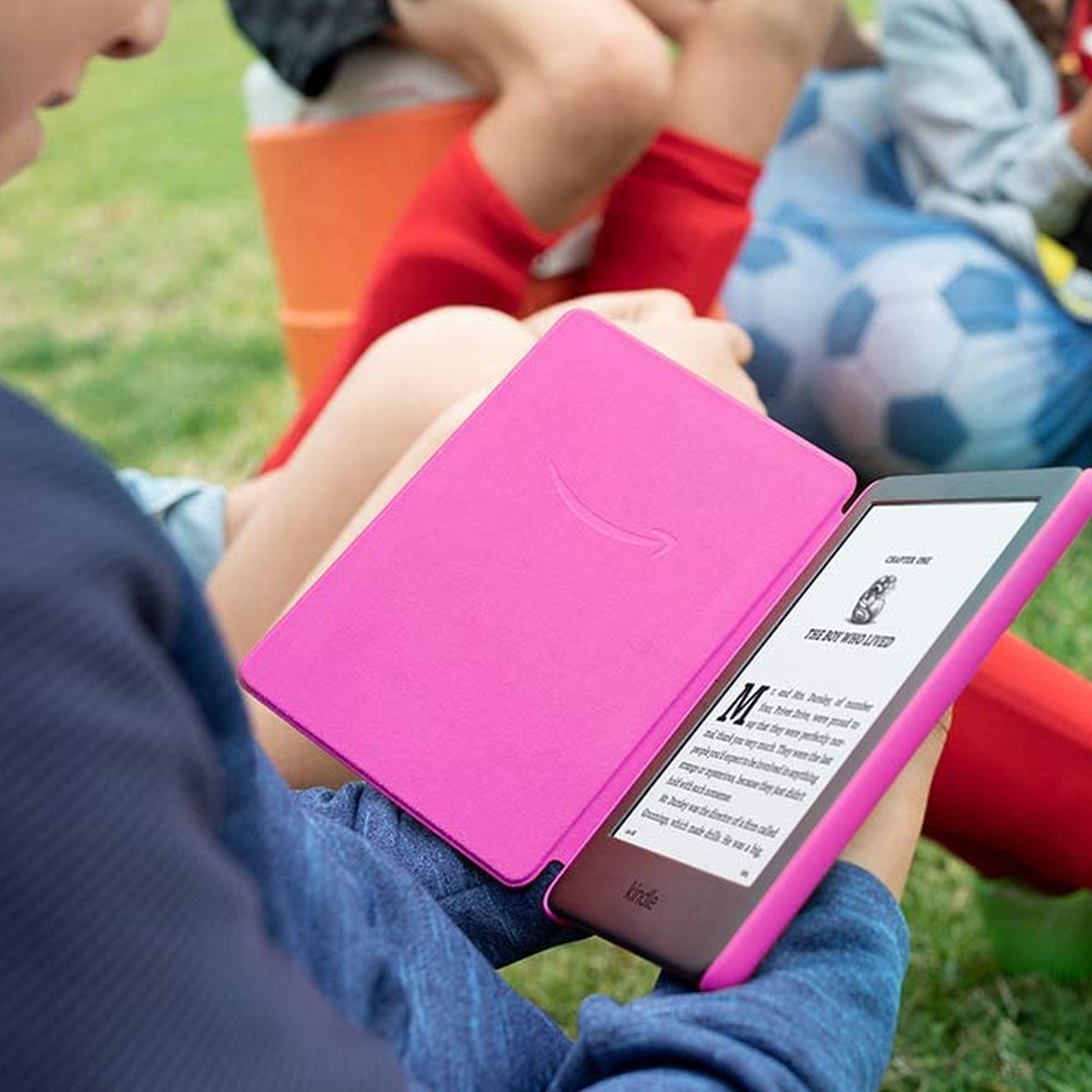 Un niño sostiene y lee un Kindle rosa mientras está sentado en el césped.