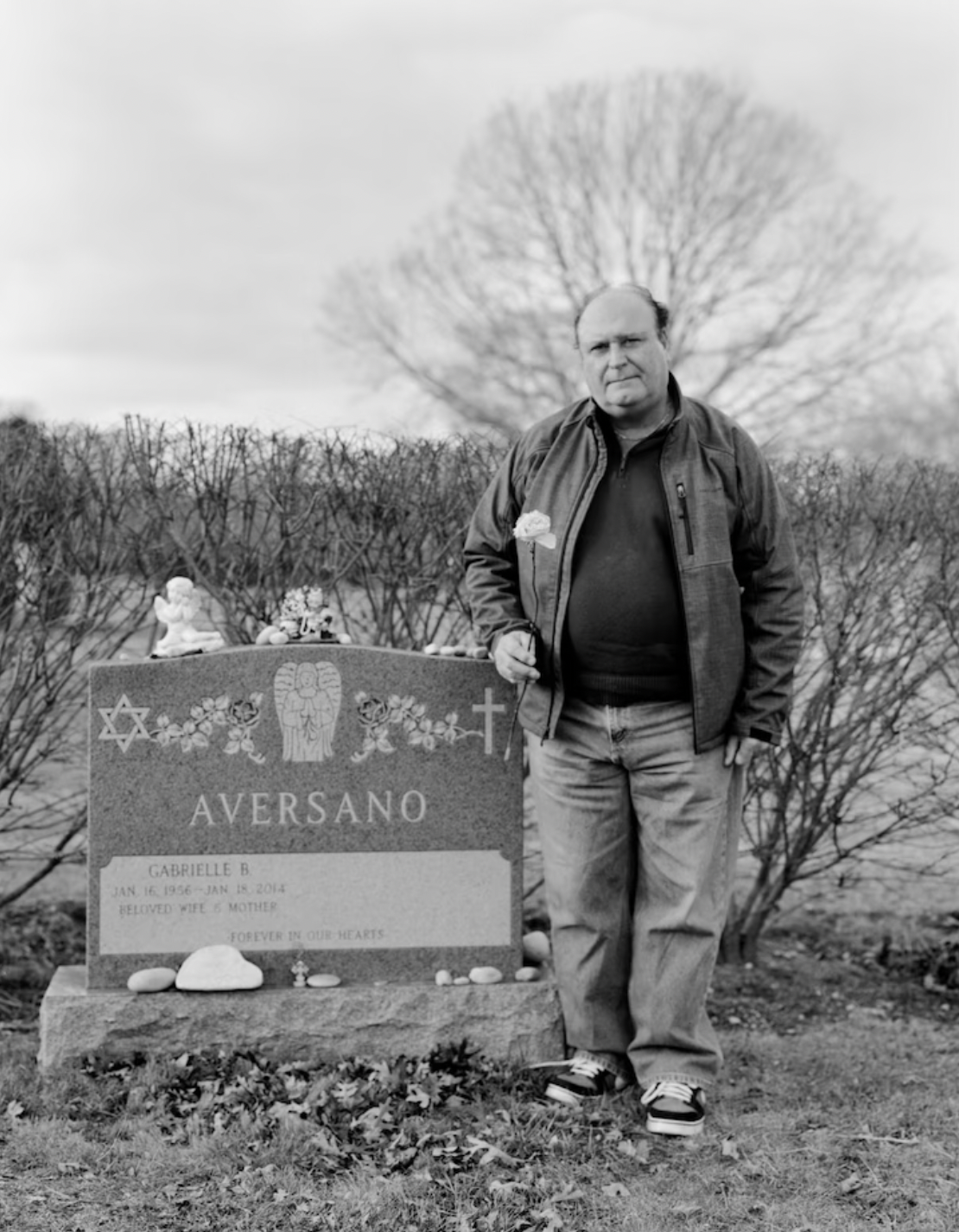 A black and white photograph of a man holding a flower by his wife's grave.  There is an open space on the tombstone where his name will be written when he dies.