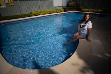 A woman in a white T-shirt and shorts, sitting on the edge of a backyard pool with her legs dangling in the water.