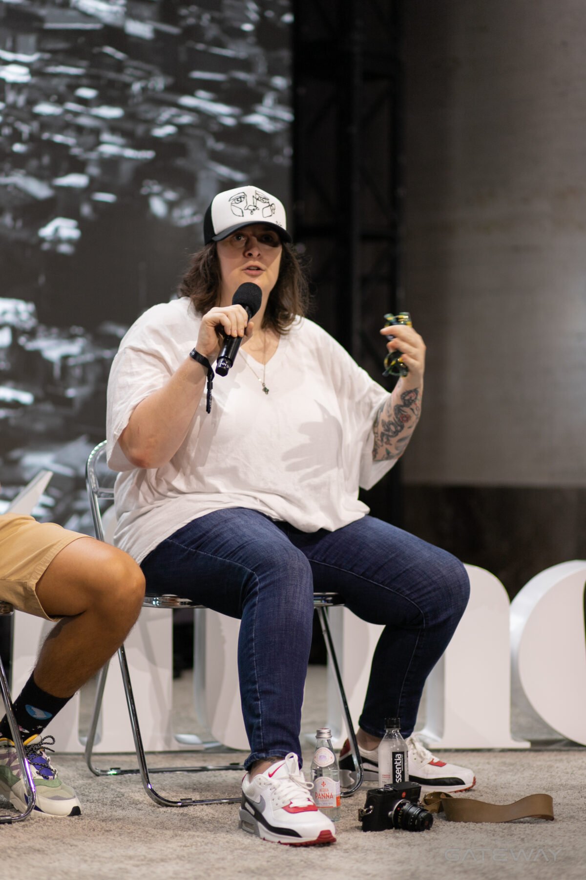 A woman in a baseball cap, white shirt, and jeans holds a microphone on a stage and gestures with her hand as she speaks. 