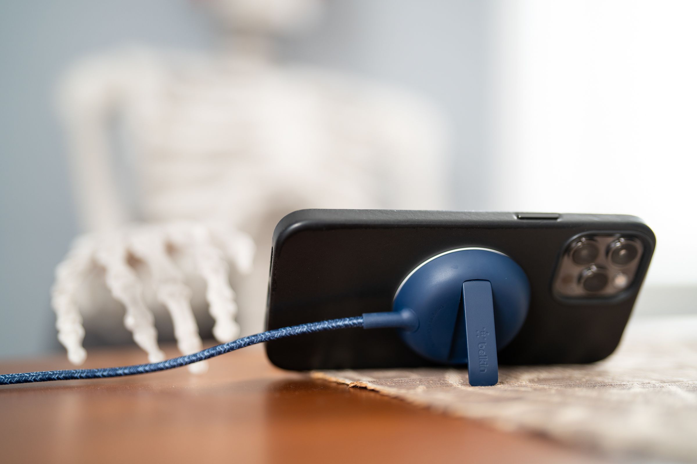 An iPhone held horizontally by a Belkin MagSafe charger holder on a table with a skeleton sitting in a chair looking at the phone.