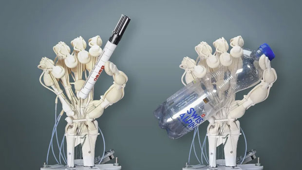 Image of two 3D printed all-in-one robotic hands, one holding a marker pen and the other clutching an empty clear plastic water bottle.  Both are placed on pedestals in front of a dark gray background.