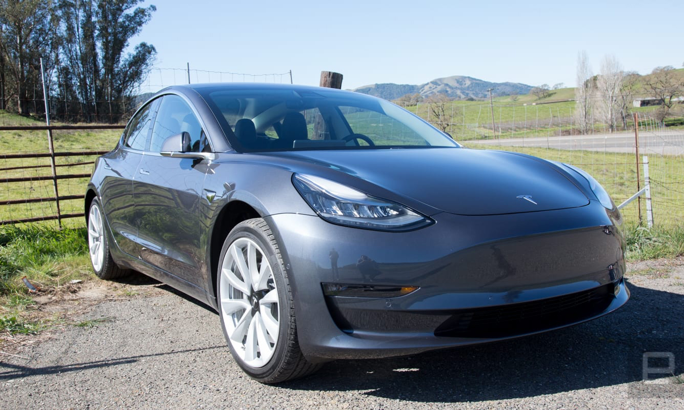 Photo of the Tesla Model 3 outdoors next to a field.  In the distance you can see green grass, trees and hills.