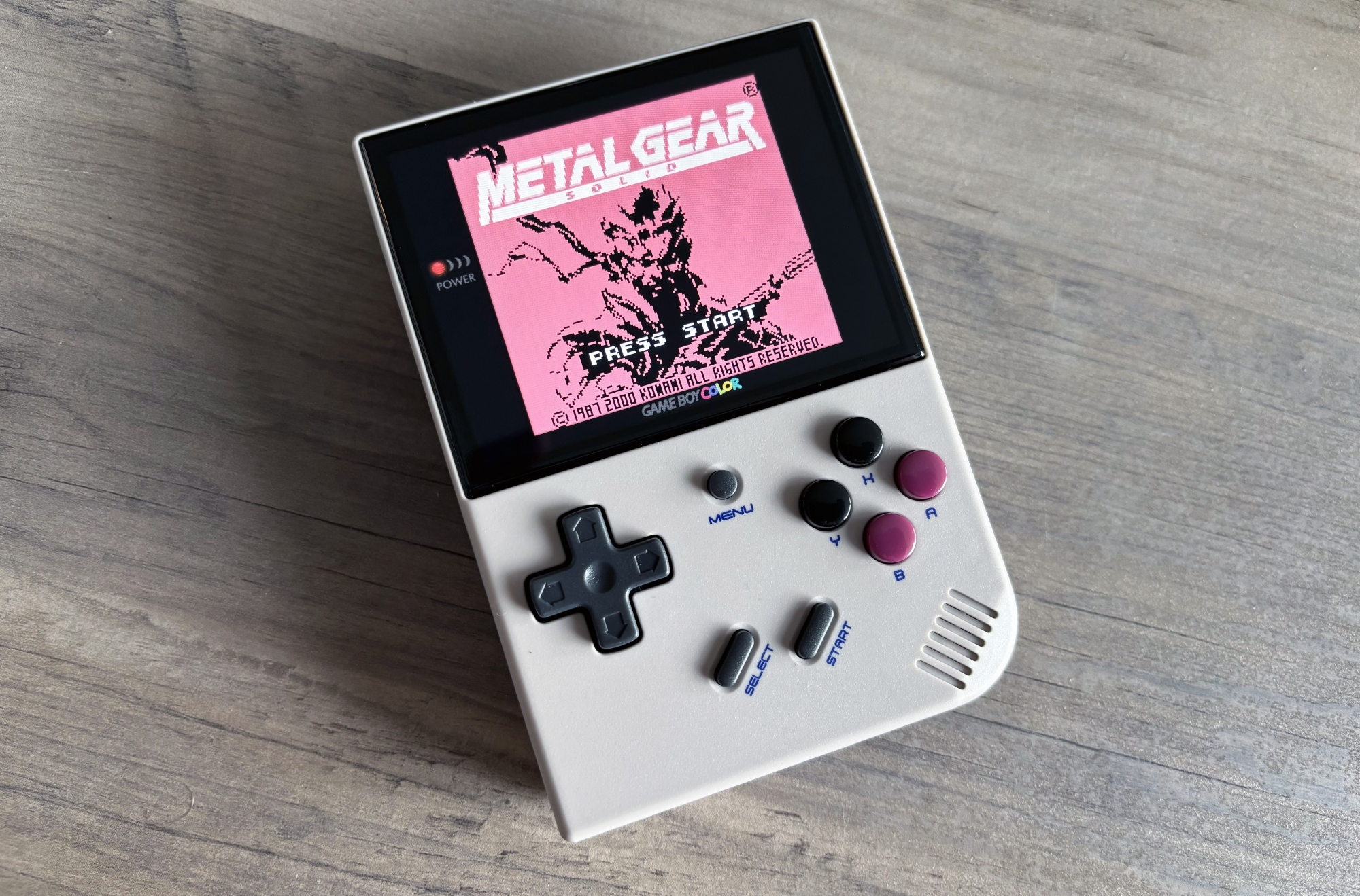 A small gaming handheld that looks reminiscent to the original Nintendo Game Boy called the Anbernic RG35XX Plus rests at an angle on a light brown wooden table. The display is turned on and showcases the start screen from the Game Boy game Metal Gear Solid.