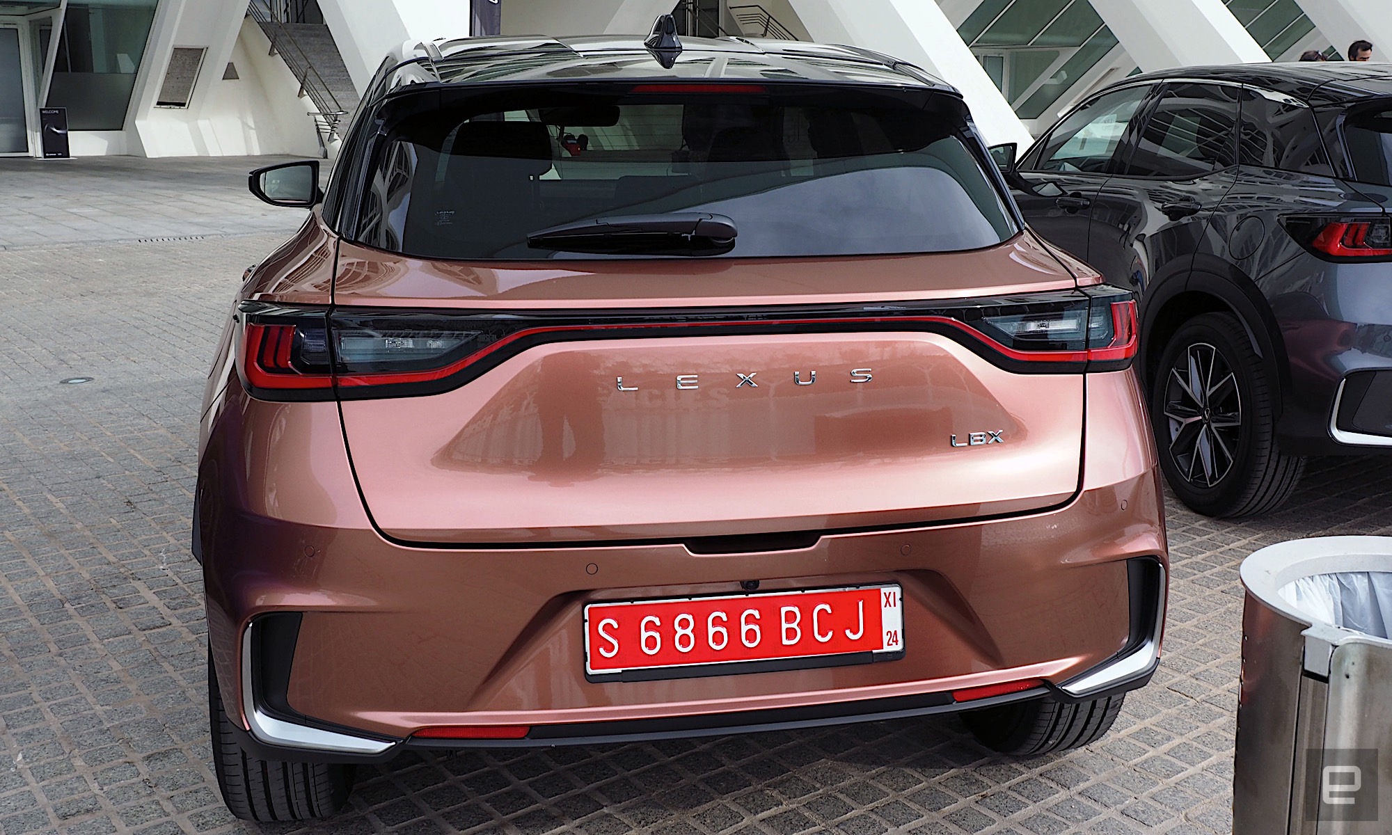 Image of the trunk of a Lexus LBX parked under the marquee of the City of Arts and Sciences in Valencia.  The car is painted in “Sonic Copper”, a kind of metallic orange.
