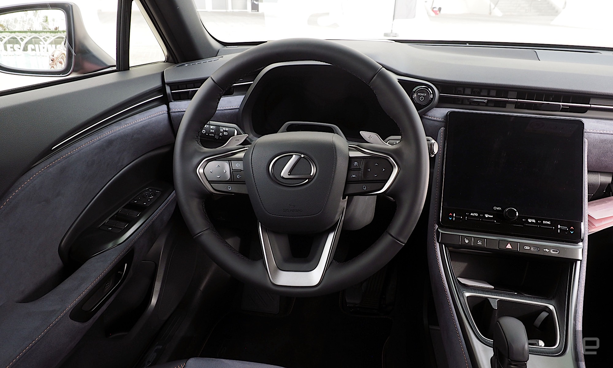 Image of the driver's position inside a Lexus LBX with a dark gray and black interior, the steering wheel is located in front of a digital instrument binnacle while an infotainment system is located to its right.