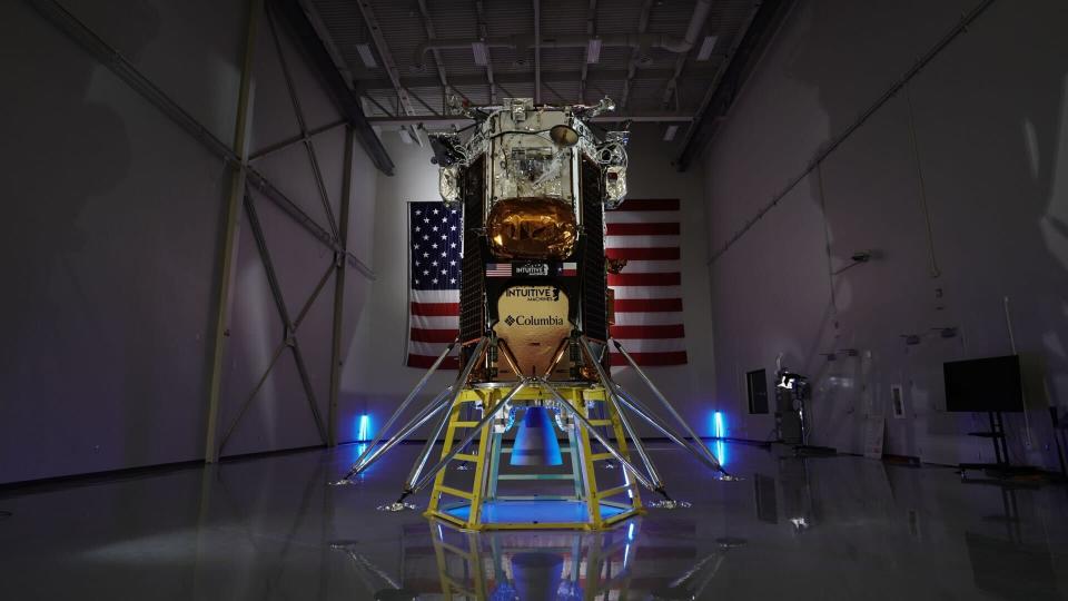 Intuitive Machines' Odysseus Nova-C lander is shown in front of an American flag in a dimly lit warehouse room.