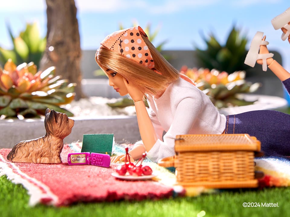 A Barbie is shown lying on a picnic table with a plastic dog in front of her and a pink phone open on her side.