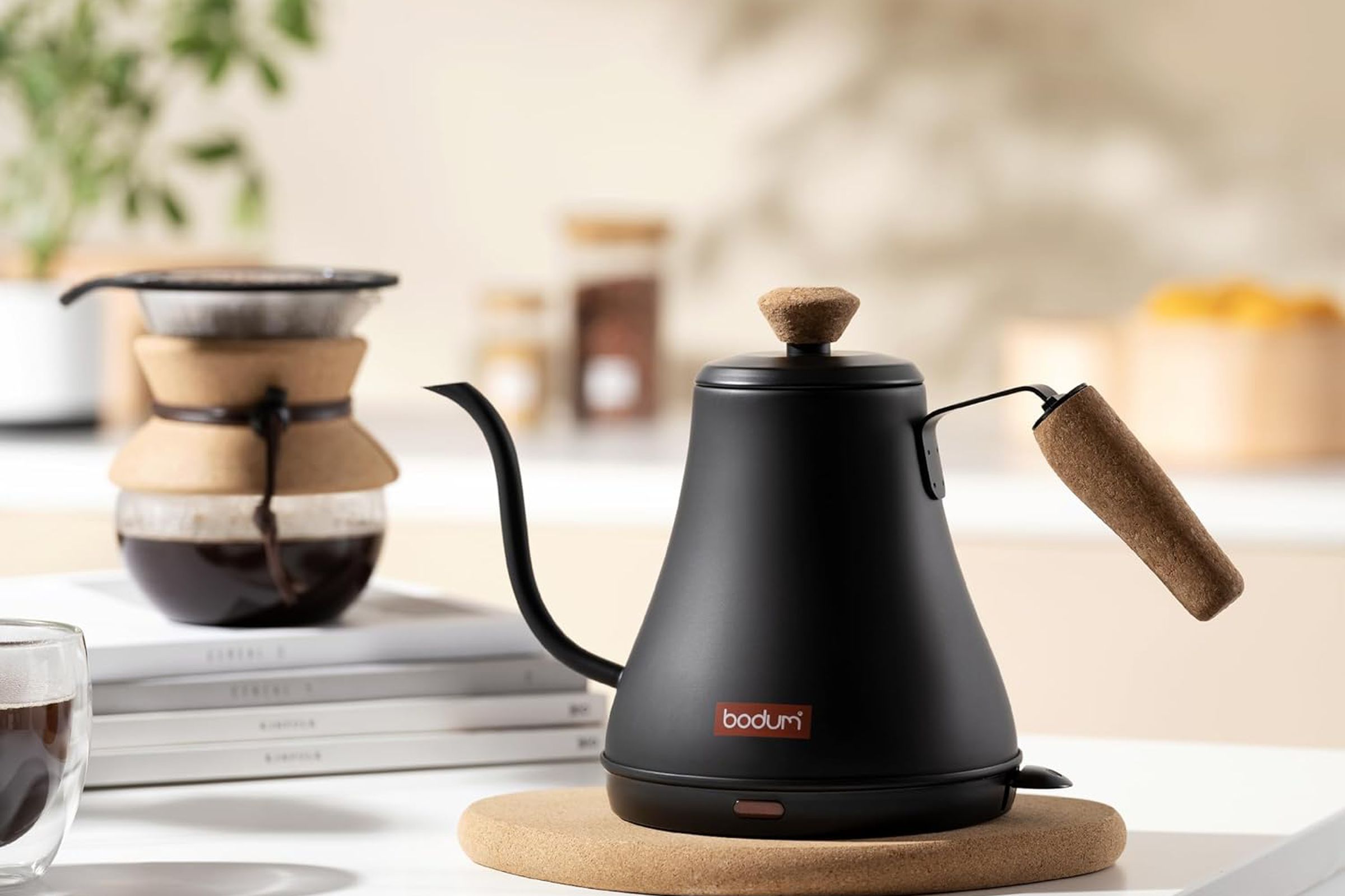 Black gooseneck electric kettle on a table, a pot of coffee in the background.