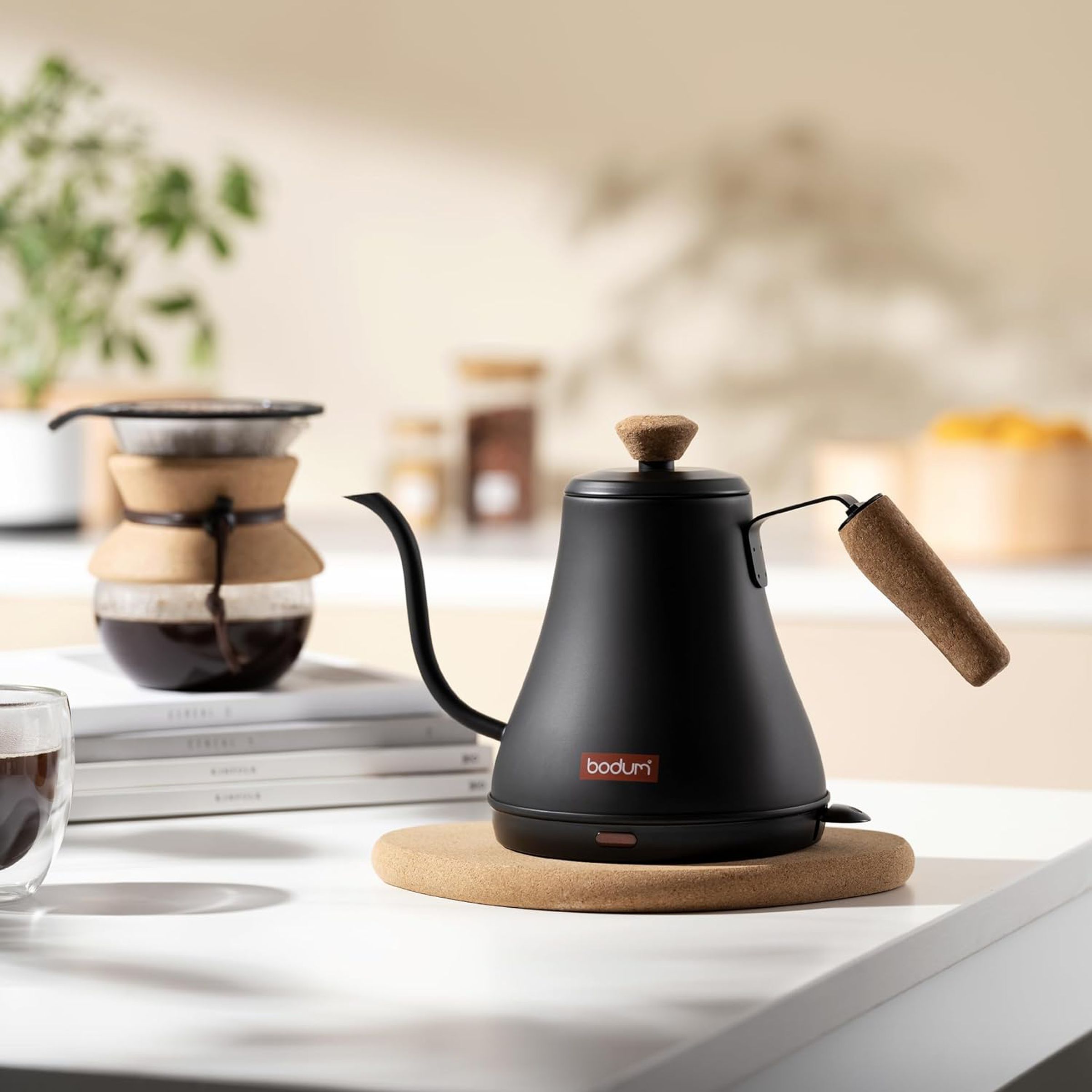 Black gooseneck electric kettle on a table, a pot of coffee in the background.