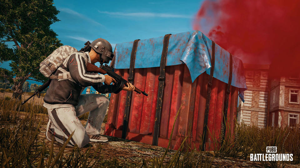 Still from PUBG, showing a player taking cover behind a box with an assault rifle.  Rural scene with an old house behind.