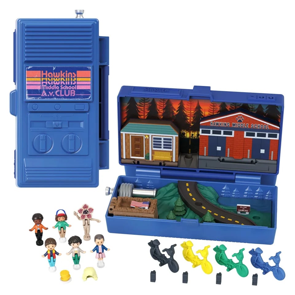 The Stranger Things Polly Pocket set and included figures are shown against a white background. It includes a compact blue walkie-talkie shown closed on the left and open on the right. The open side contains a depiction of Hawkins, including the high school. There are also figures of Eleven, Mike, Will, Lucas, Dustin and a Demogorgon, plus mini walkie-talkies and bikes.