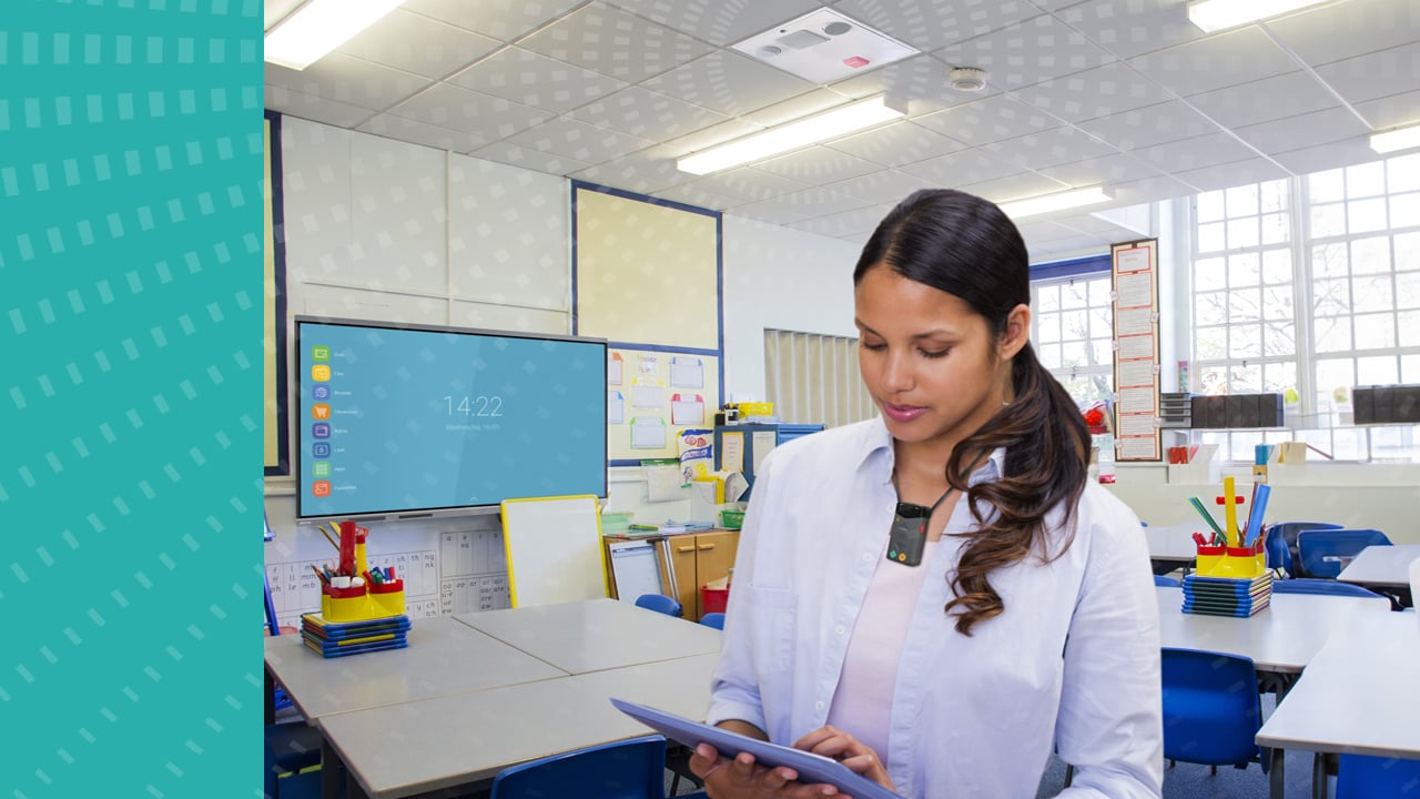 A classroom with a teacher using a FrontRow microphone and touching a tablet screen. There is an ezRoom speaker mounted in the ceiling.