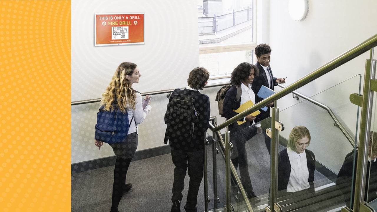 High school students walking down a hallway. The TimeSign screen displays the fire drill alert. 