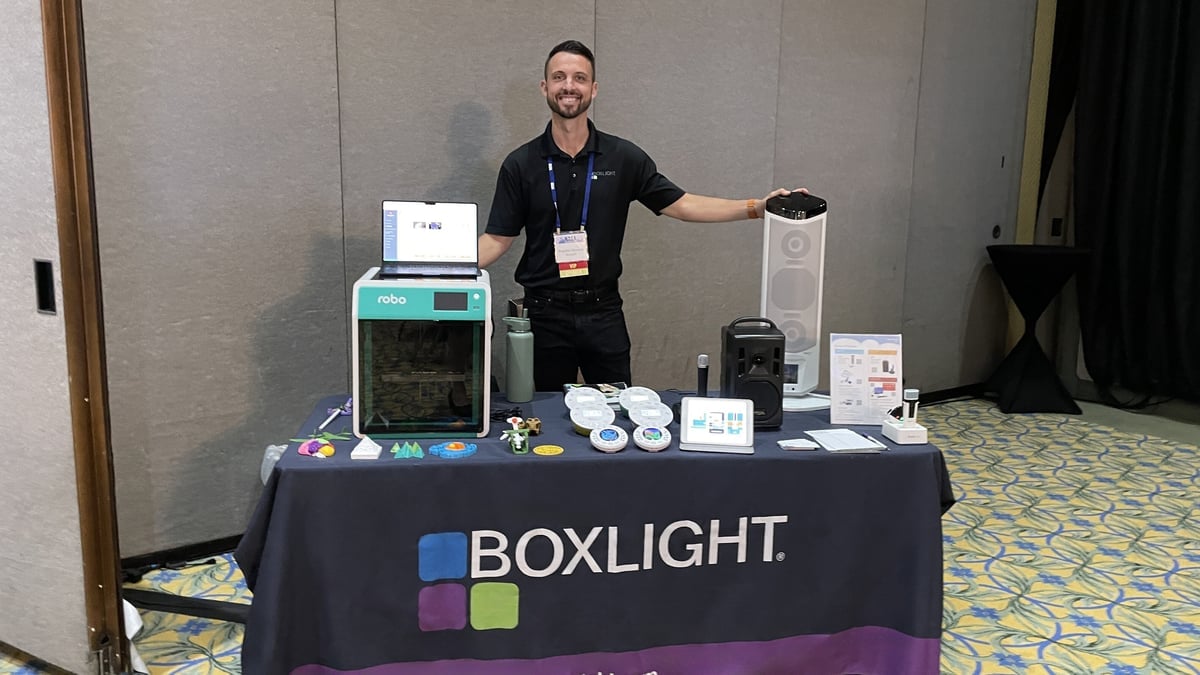 Braydon Moreno, director of STEM at Boxlight, stands behind a table covered with Boxlight solutions, including the award-winning Robo E4 3D printer that comes with the MyStemKits curriculum, a set of Labdisc all-in-one science labs, a FrontRow Lyrik portable audio system, a FrontRow Juno audio speaker tower, and Action! teacher's microphone. 