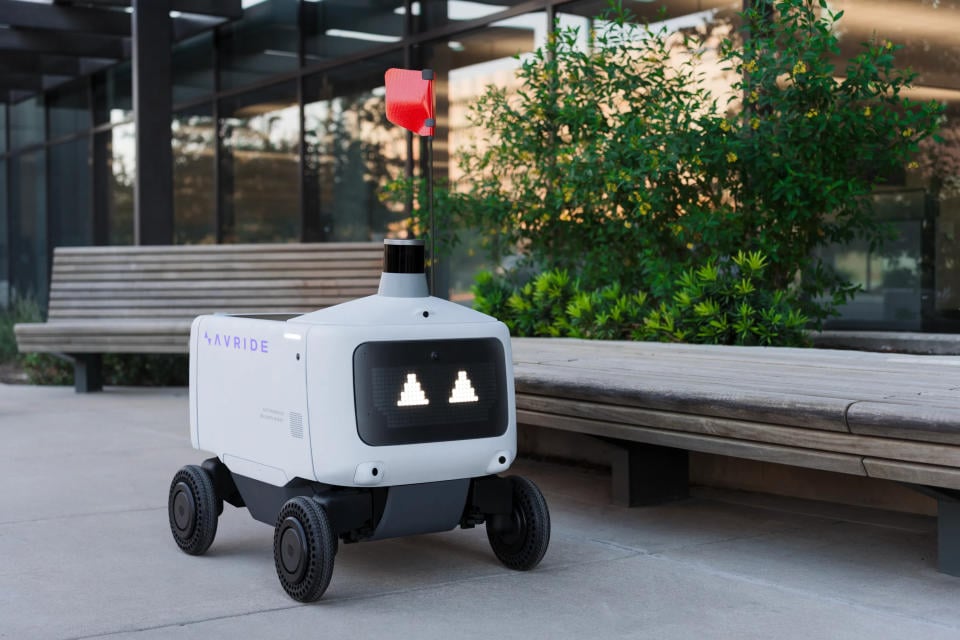 An Avride delivery robot with triangular eyes parked next to a bench.