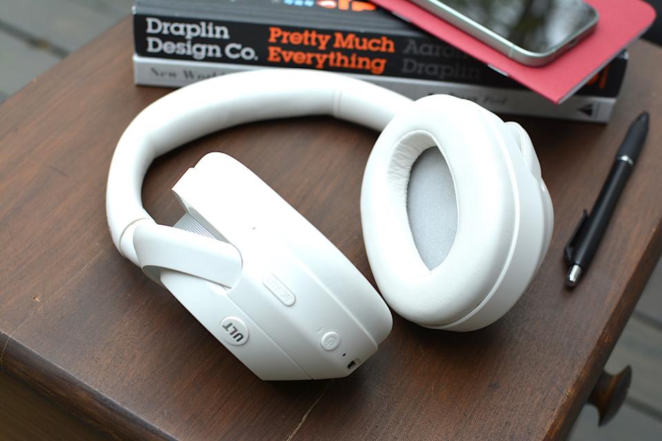 White headphones sitting on a wooden table from above with a red notebook and a phone in the background, on top of a black and white book.