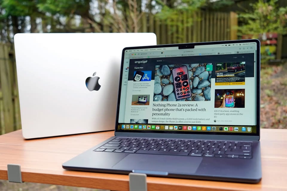 The macbook air m3 2024 sits on a wooden table outside, with a fence and some trees. 