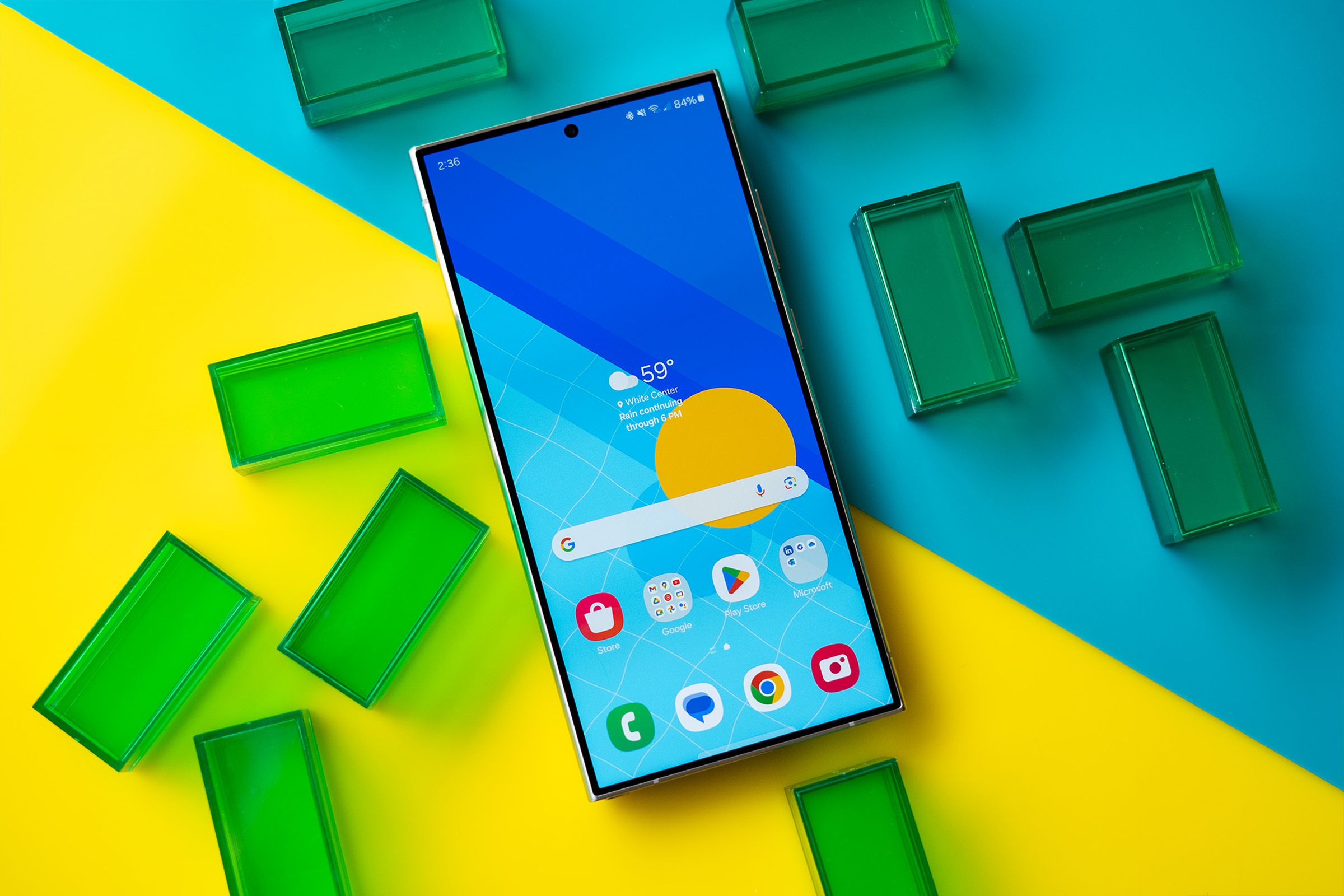 Samsung Galaxy S24 Ultra showing a blue and yellow homescreen, on a blue and yellow background with green translucent rectangles.