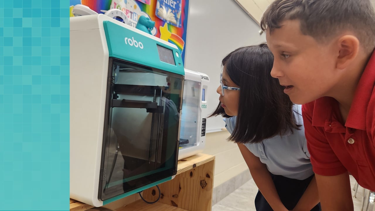 Two elementary school students watch the progress of printing their design through the glass door of the Robo E4 3D printer.