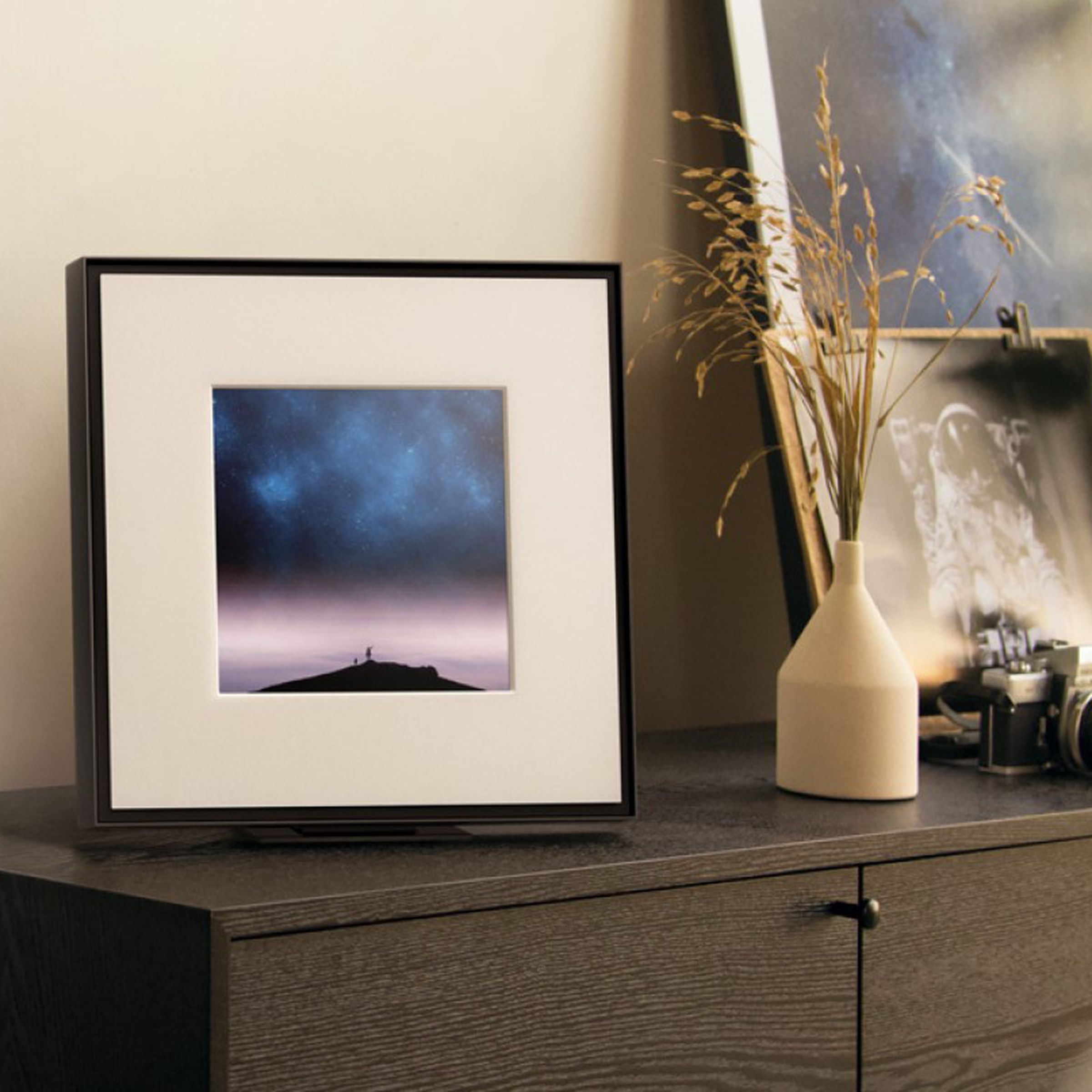 Samsung’s Music Frame speaker sitting on a brown cabinet beside a pair of cameras and some plants.