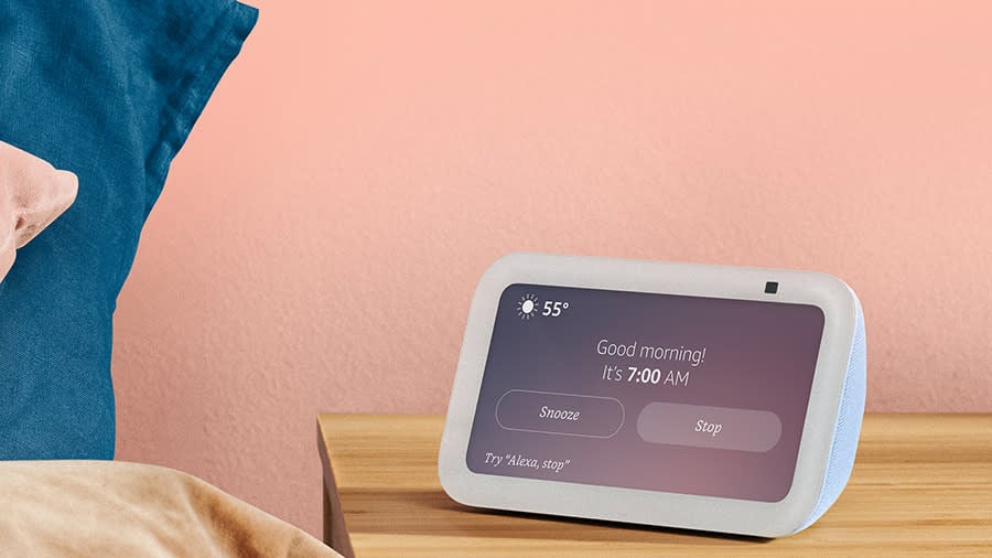 The Echo Show 5 smart display sits on a nightstand in a bright pink painted room. The screen reads 