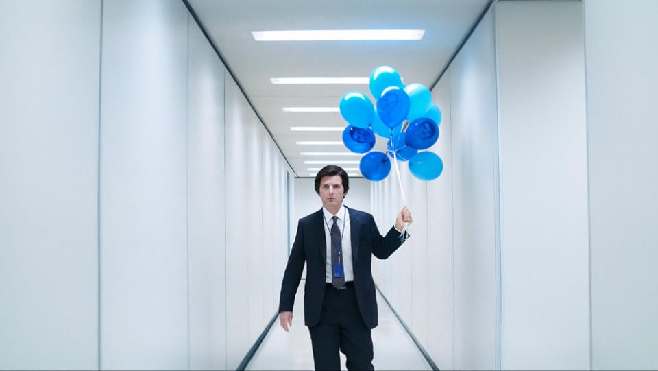 Adam Scott (wearing a suit) holds a stack of blues in a white hallway in Severance.