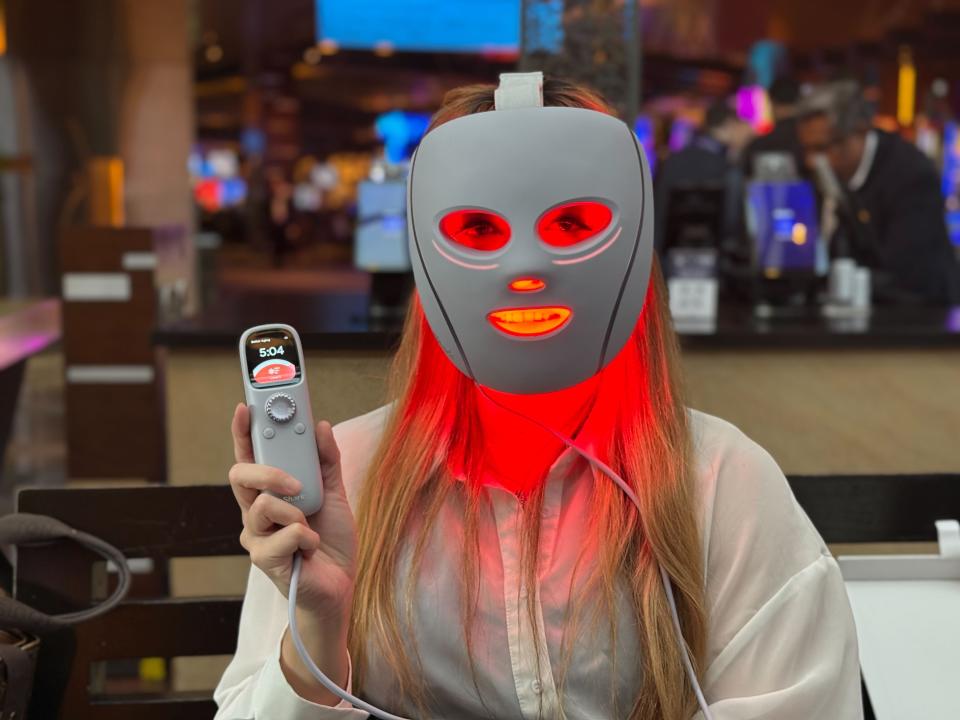 A woman wearing the Shark CryoGlow mask, which glows with red light coming from its holes. The woman also holds a remote control with a dial and a small screen.