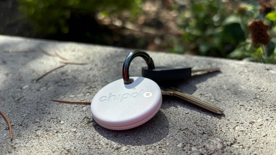 The Chipolo One Point tracker is on a keychain with a pair of keys and rests on a concrete surface with plants in the background. 